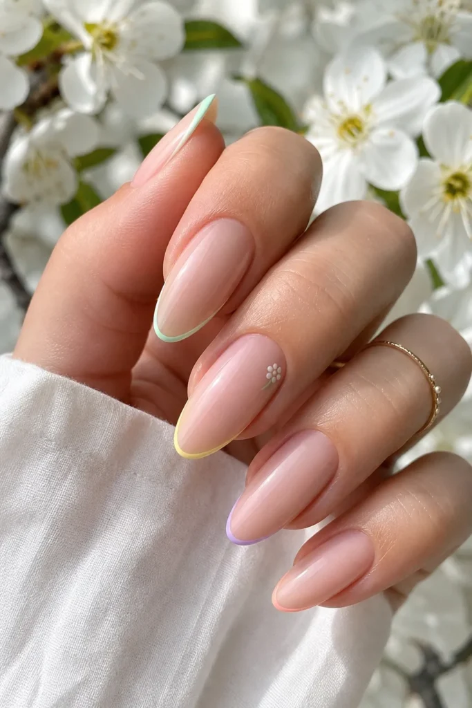 A close-up photograph of a hand with elegant, long oval-shaped nails featuring a 'Baby’s Breath' micro-French tip design. Each nail has a sheer, glossy nude-pink base with an ultra-thin stroke of a different pastel color—mint green, butter yellow, lavender, and peach—along the very edge. One nail includes a tiny, minimalist white flower detail near the cuticle, set against a bright, airy background of white spring blossoms.
