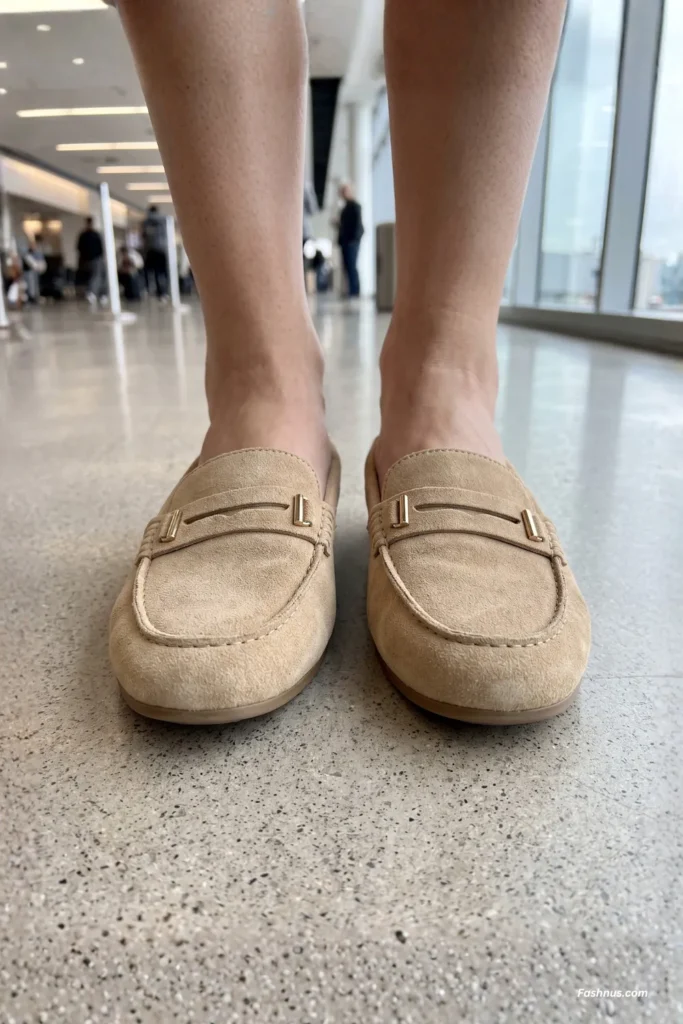 Front close-up view of a young woman's feet wearing beige suede loafers with minimalist gold metal accents on a light gray speckled floor.