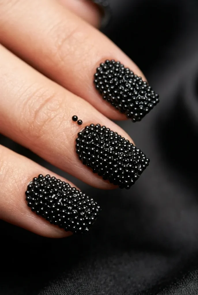 A tight close-up of fingernails completely covered in tiny, spherical glossy black micro-beads, creating a textured caviar look.