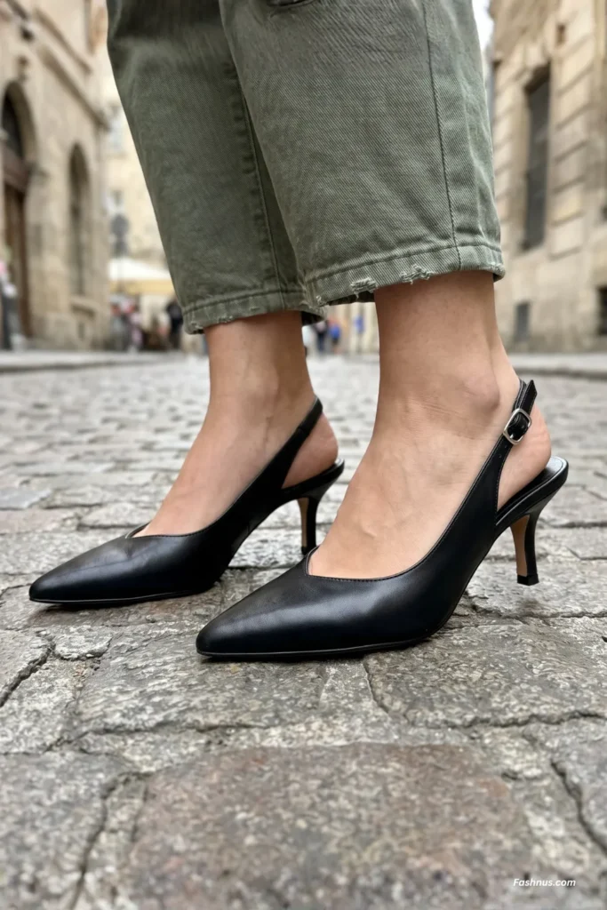 A young woman standing on a cobblestone street wearing pointed-toe black leather slingback kitten heels paired with green utility trousers.