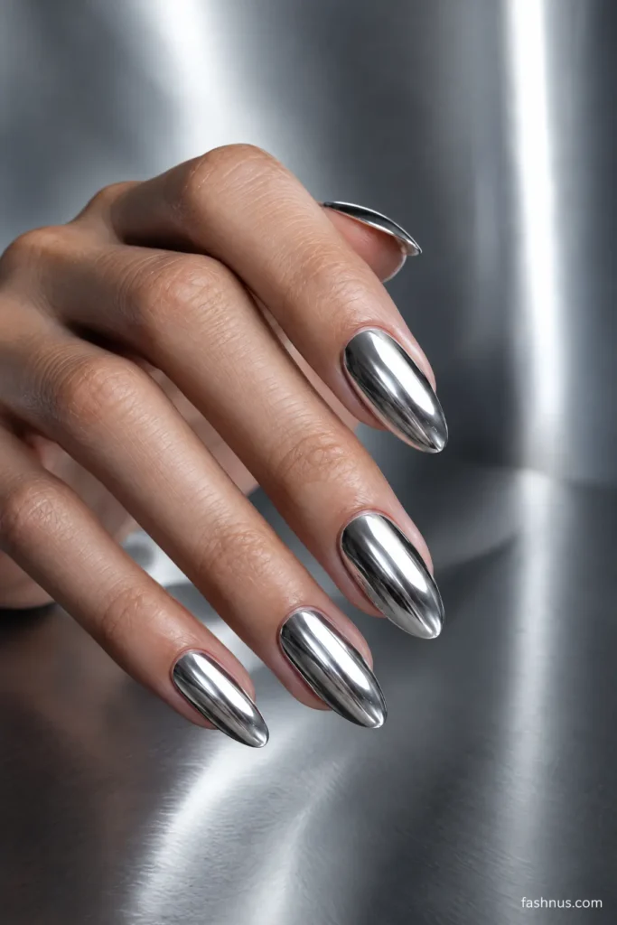 A young Western woman's hand featuring glossy silver chrome almond-shaped nails with a reflective, futuristic finish against a cool-gray background.