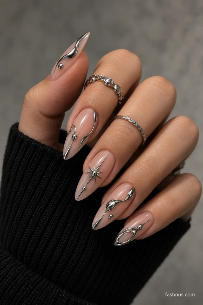 A young Western woman's hand with long normal-pink almond-shaped nails featuring raised silver chrome curves, metallic droplets, a celestial starburst, and abstract oval details, complemented by silver rings and a black knitted sleeve.