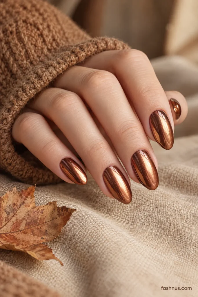A young Western woman's hand showcasing glossy copper metallic almond-shaped nails beside autumn leaves and warm beige fabric.