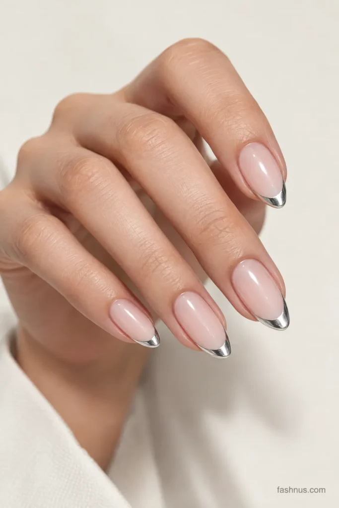 A young Western woman's hand featuring elegant nude-pink nails with thin reflective silver French tips against a soft ivory background.