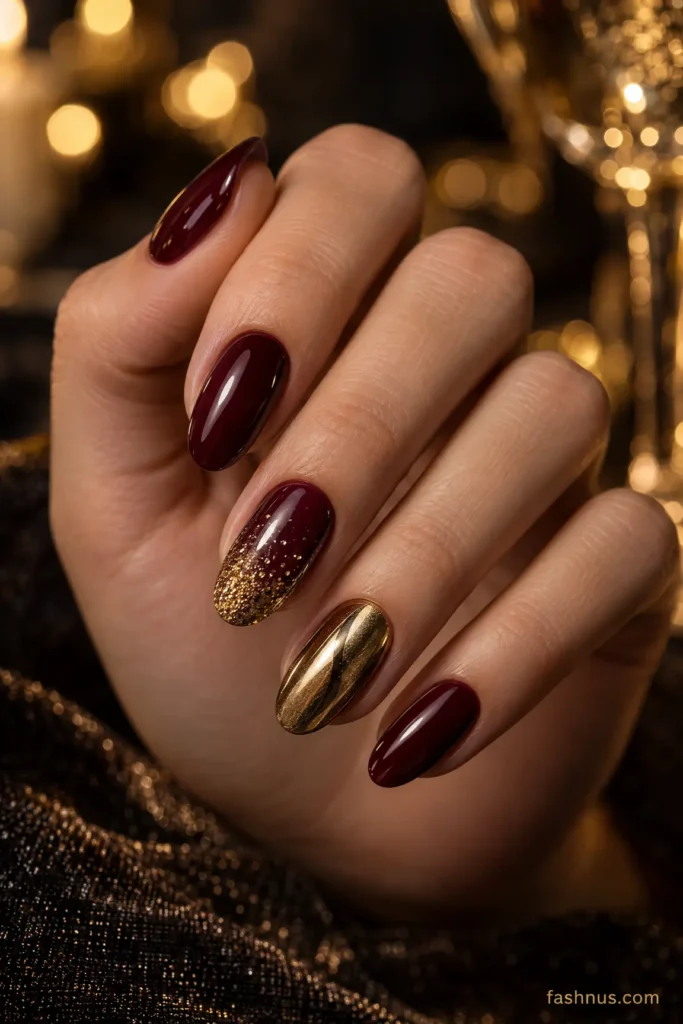 A young Western woman's hand displaying deep burgundy nails with sparkling gold glitter gradients and glossy metallic accents against a softly illuminated dark background.